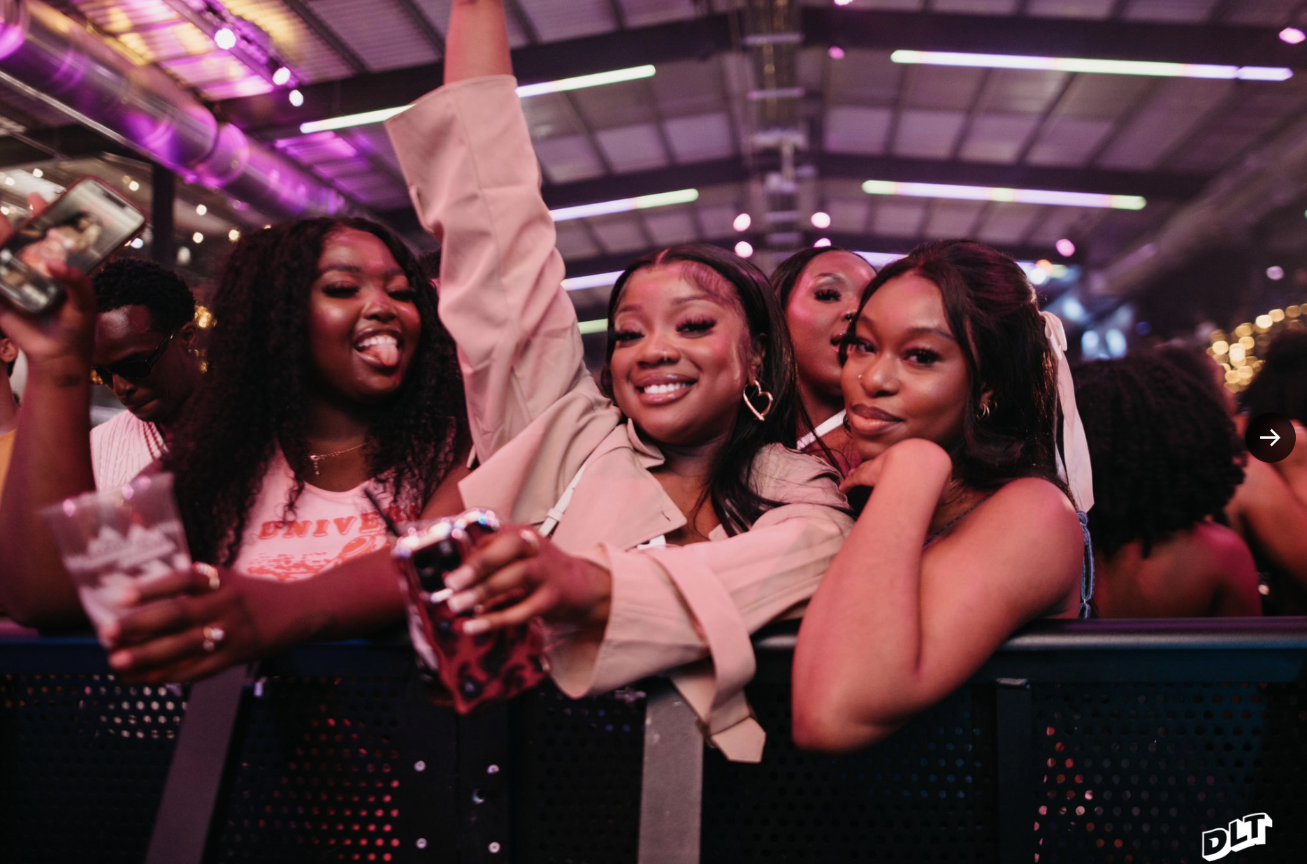 Group of three women smiling and posing for a photo at a lively party or event. One woman has her arm raised. The background features a party atmosphere