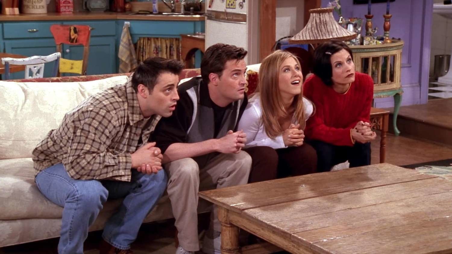 Joey Tribbiani, Chandler Bing, Rachel Green, and Monica Geller are sitting on a sofa, leaning forward, intently watching something off-screen