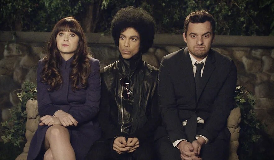 Zooey Deschanel, Prince, and Jake Johnson sit side by side on a bench, each with distinct expressions. Zooey is in a coat, Prince in a leather jacket, Jake in a suit