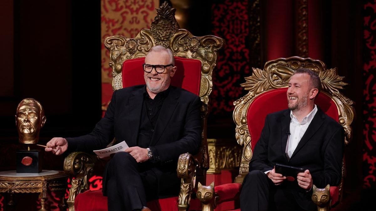 Two men, both seated on ornate chairs. The man on the left holds a paper, smiles broadly, and wears a black suit. The man on the right, also smiling, wears a dark suit and holds a tablet