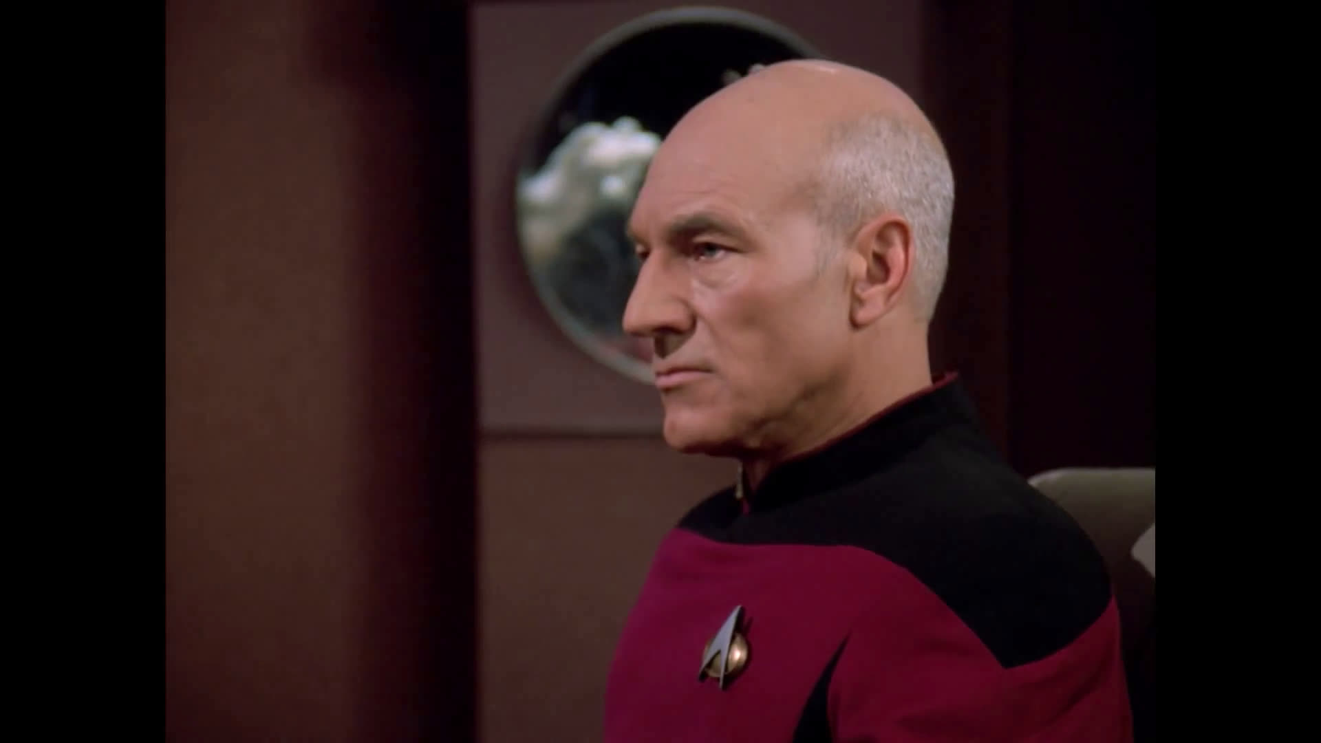 Patrick Stewart, as Captain Jean-Luc Picard from Star Trek, wearing a Starfleet uniform with a communicator badge, appears serious while seated