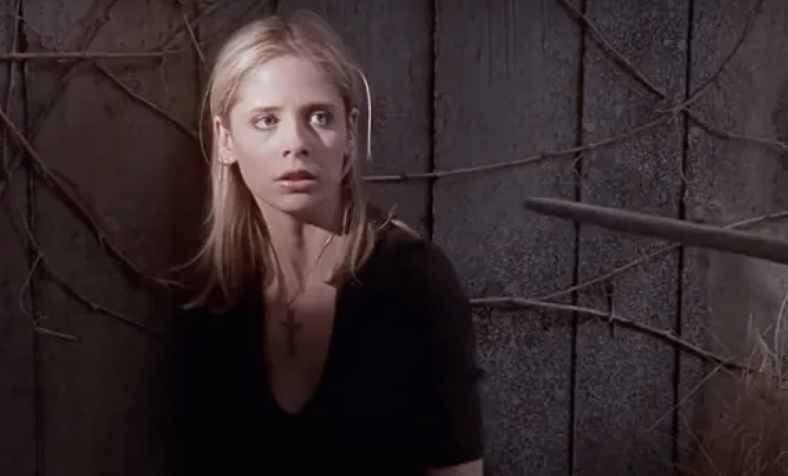 Sarah Michelle Gellar, in a scene from Buffy the Vampire Slayer, wears a black top and a cross necklace while looking to the side. A wooden stake points towards her