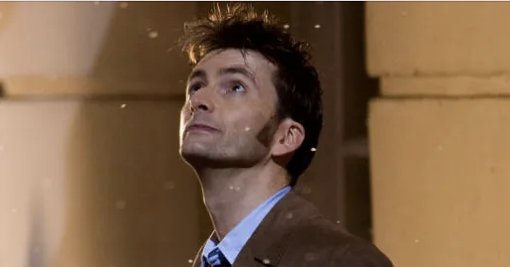 David Tennant, dressed in a suit and overcoat, looks up with a pensive expression, with snow falling around him