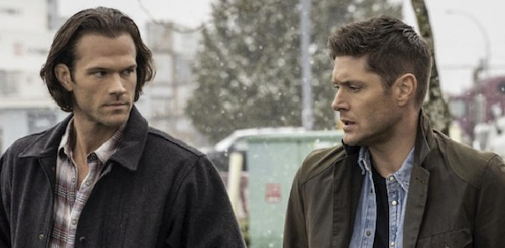 Jared Padalecki and Jensen Ackles stand outdoors in casual jackets, having an earnest conversation. Snow is falling lightly in the background