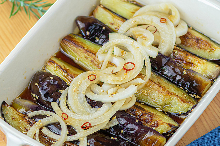 白いオーブン皿に盛られた焼きナスの上に、スライスされた玉ねぎと調味料がかけられている料理。