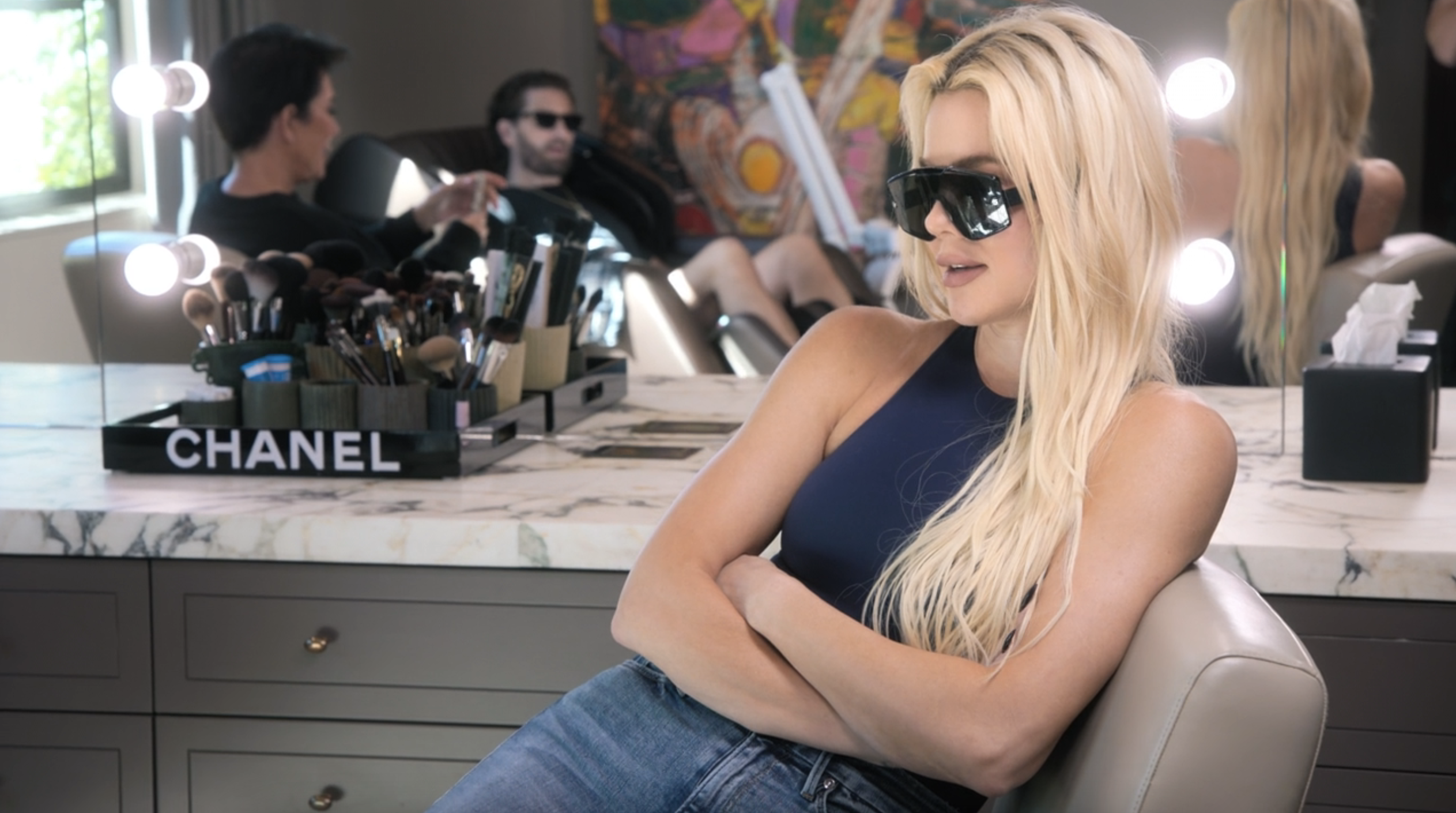 Kim Kardashian and Kanye West in a makeup room, Kim is in a blue top and jeans, wearing large sunglasses and seated, with makeup tools on the counter