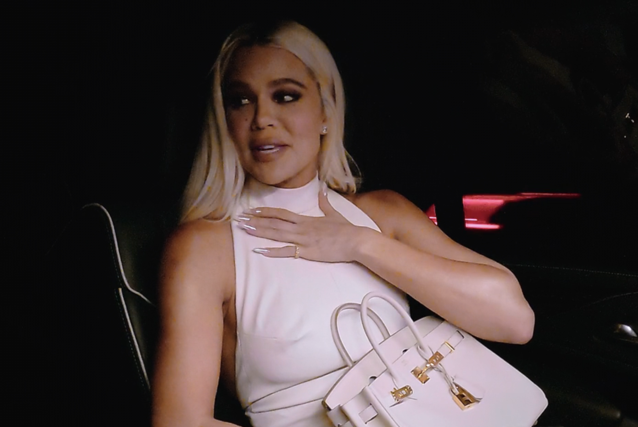 Khloé Kardashian sits in a car wearing a sleeveless white top, holding a white handbag, and looking to the side with one hand on her chest