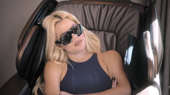 Khloé Kardashian is seated in a massage chair with sunglasses on, appearing relaxed. She is wearing a sleeveless top and her arms are resting on the chair