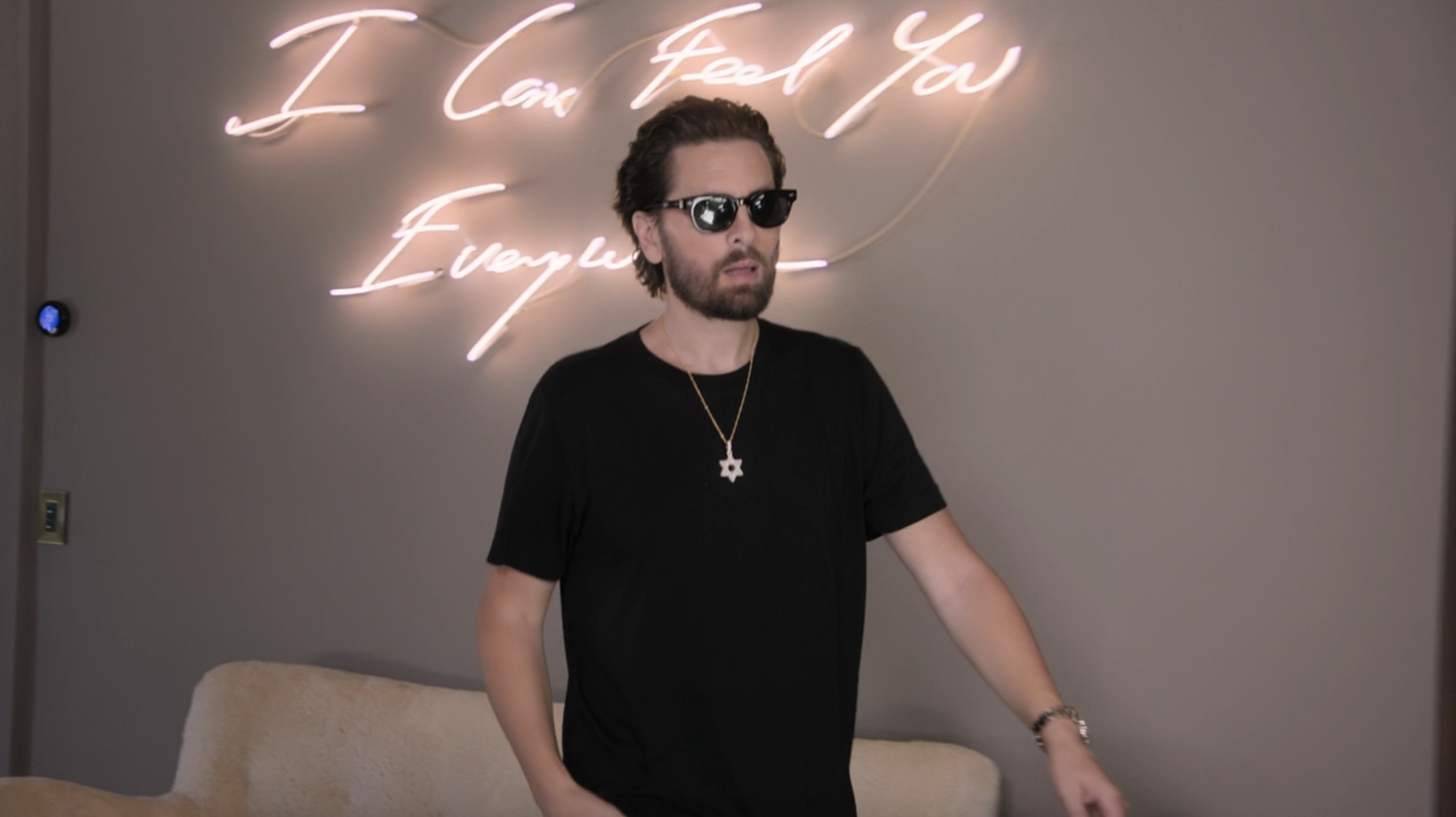 Scott Disick wearing sunglasses and a black t-shirt stands in front of a neon sign that reads "I Can Feel You Everywhere."