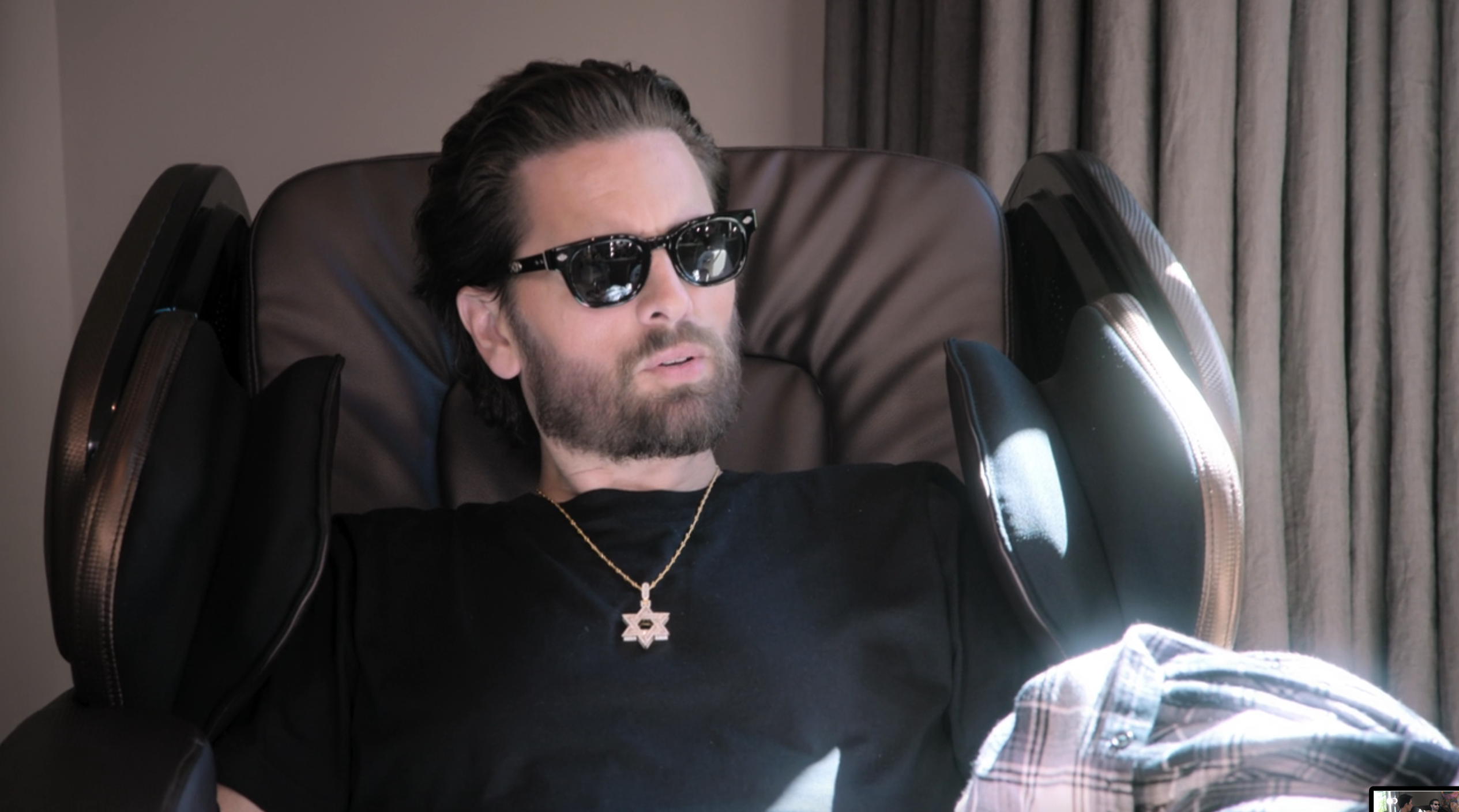 A man with dark sunglasses and a black t-shirt is sitting in a massage chair. He wears a Star of David necklace