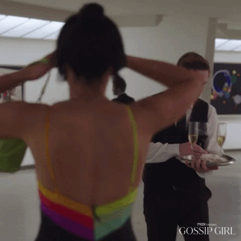 A person with their back to the camera adjusts their hair while another person holding a tray with drinks approaches. Scene from Max Original's Gossip Girl