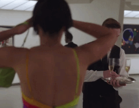 A person with their back to the camera adjusts their hair while another person holding a tray with drinks approaches. Scene from Max Original's Gossip Girl