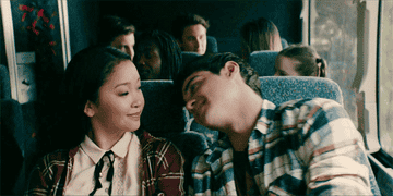 Lana Condor and Noah Centineo share a sweet moment on a bus, smiling and looking at each other. The bus is filled with other passengers in the background