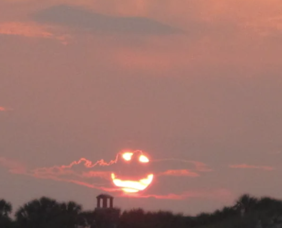 Sunset with clouds forming a smiling face, resembling an emoji. Palms and a small structure are silhouetted in the foreground