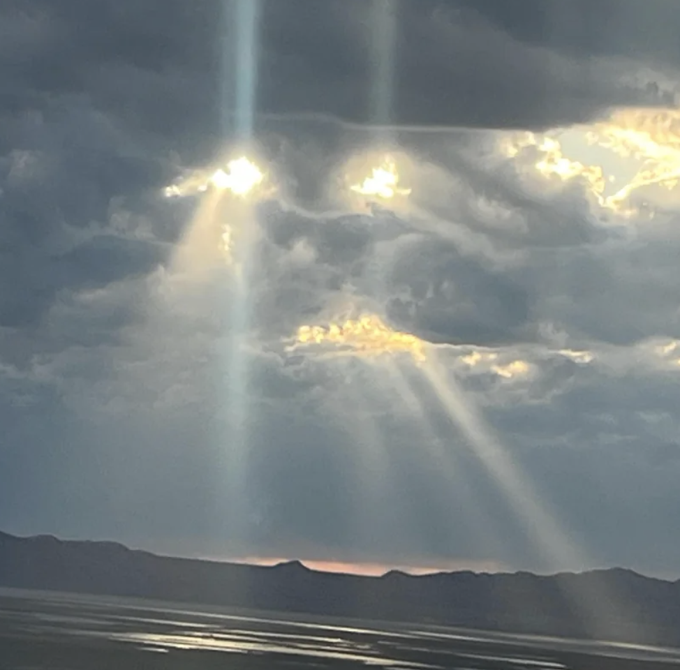 Sunbeams break through cloudy skies, casting light over a serene landscape with distant mountains and water