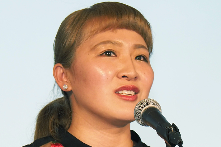 壇上でスピーチをしている女性。シースルー素材と花柄デザインのドレスを着用。