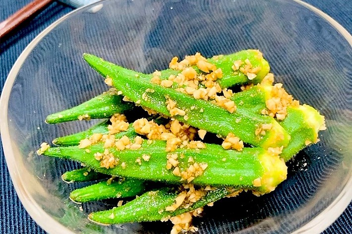 透明なガラスの器に盛られたオクラが、胡麻と調味料で和えられている。器は木製のテーブルに置かれている。