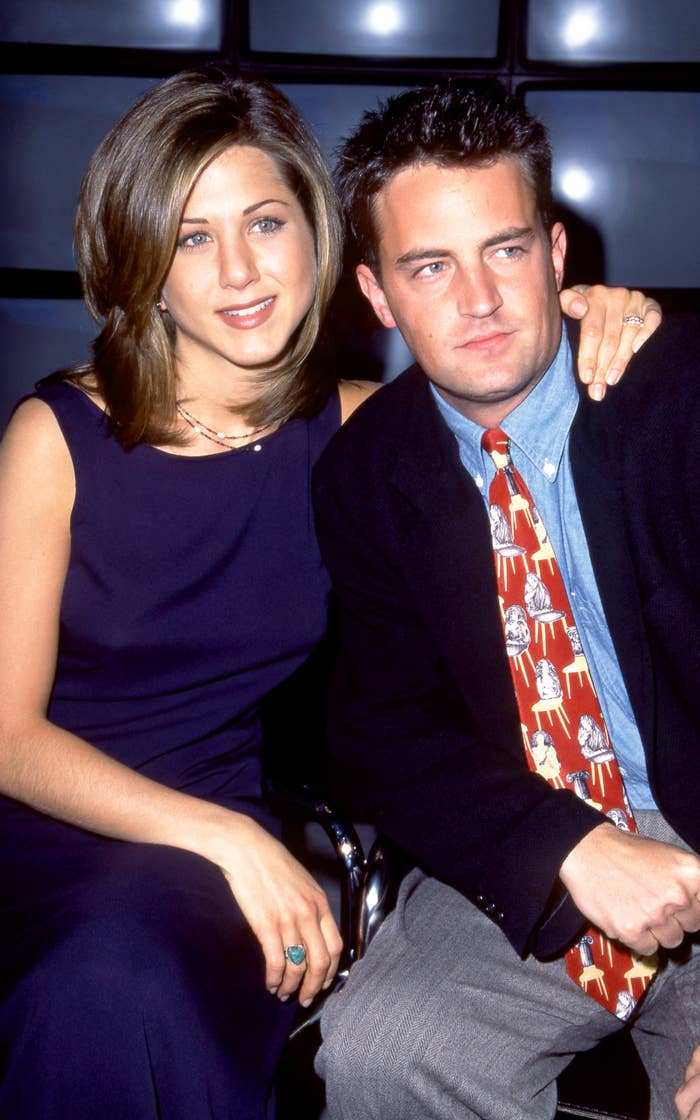 Jennifer Aniston in a sleeveless dress sits next to Matthew Perry in a suit and colorful tie, with Jennifer's arm around Matthew's shoulder, both smiling