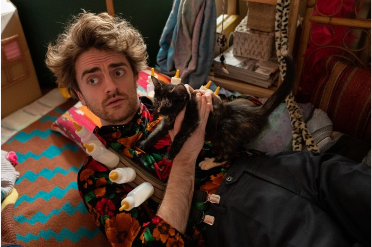 A person lying on a bed wearing a floral shirt and holding a black and white kitten. They have a surprised expression and several baby bottles attached to their shirt
