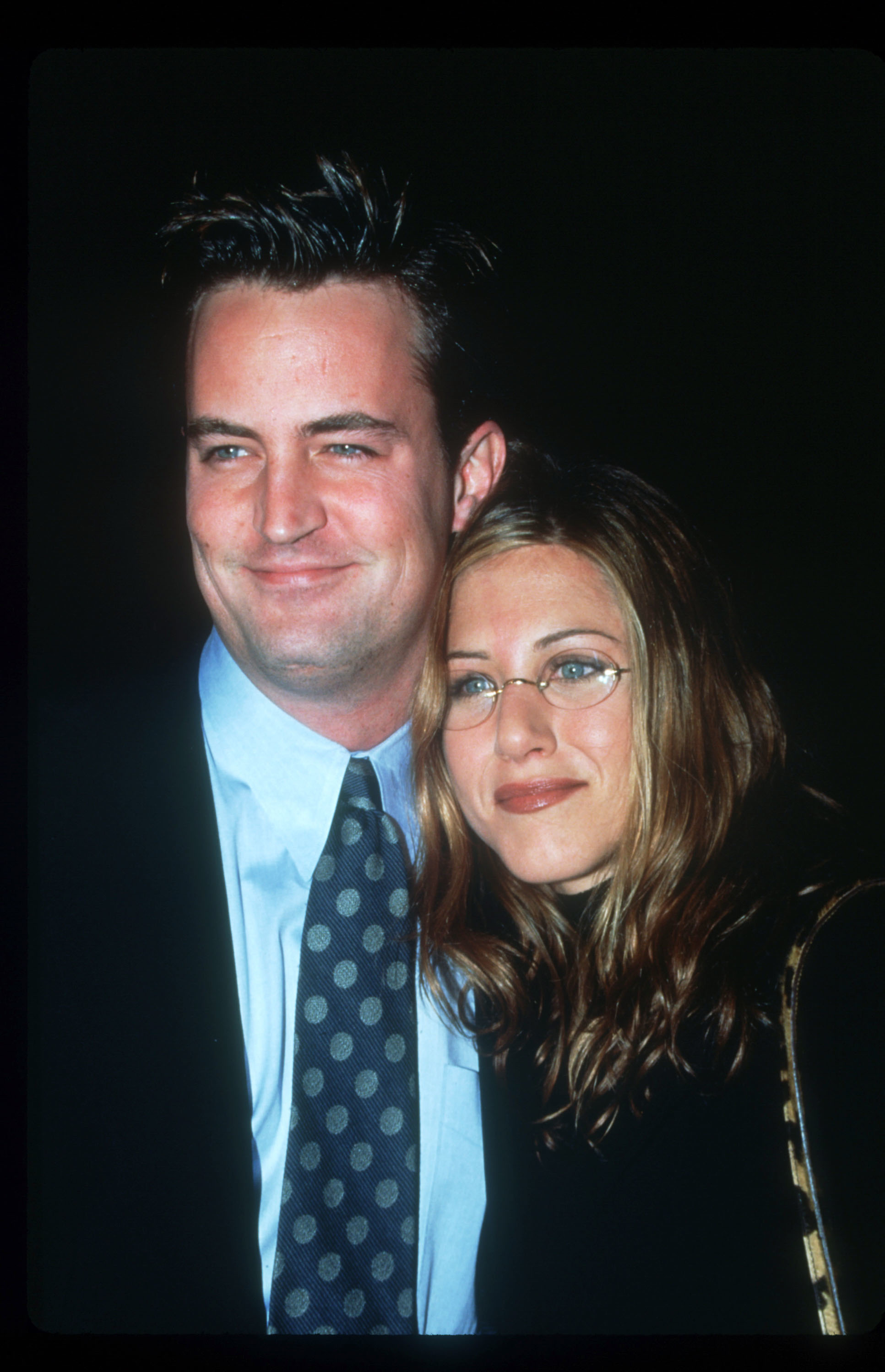 Matthew Perry and Jennifer Aniston, with him in a suit and tie, and her in glasses, smiling together