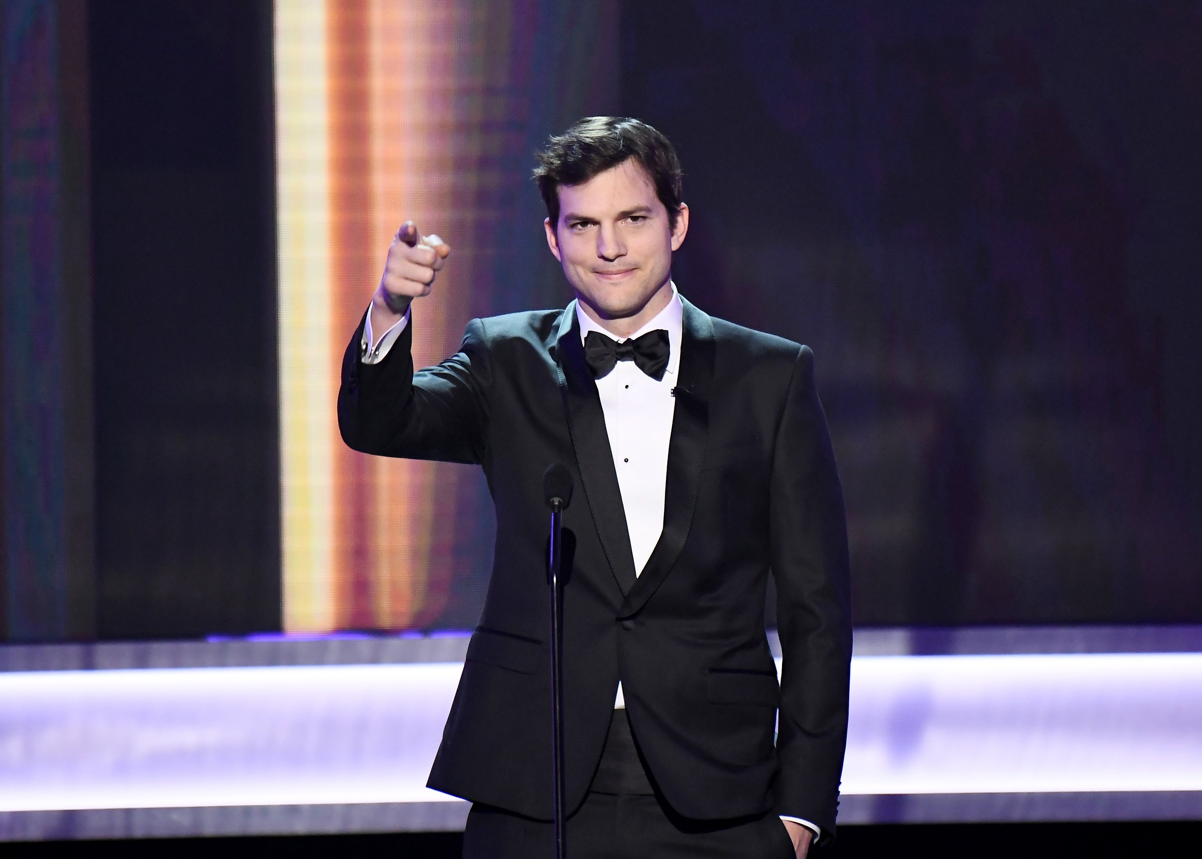 Ashton Kutcher in a black tuxedo with a bow tie, standing on stage pointing toward the audience