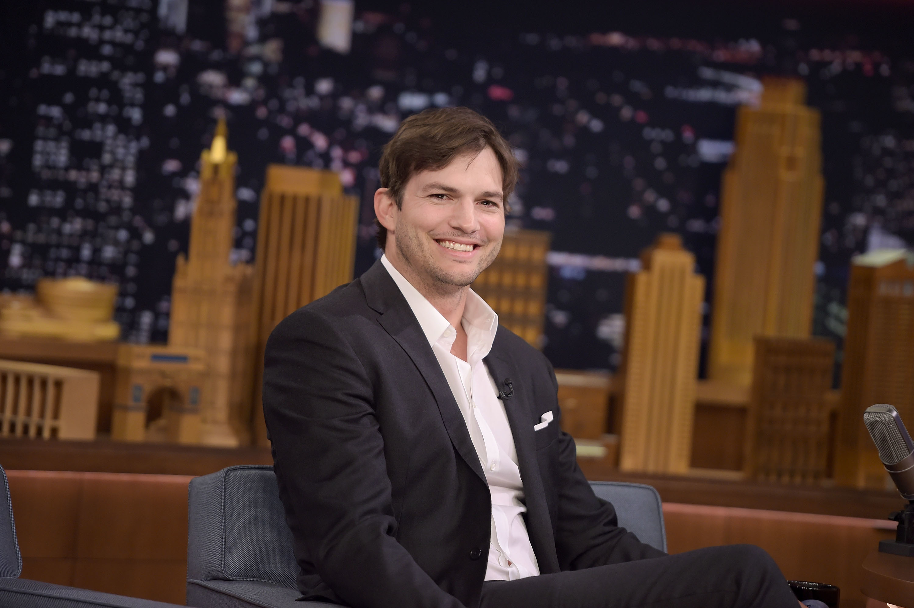 Ashton Kutcher smiles while seated on a late-night talk show, wearing a blazer over a dress shirt