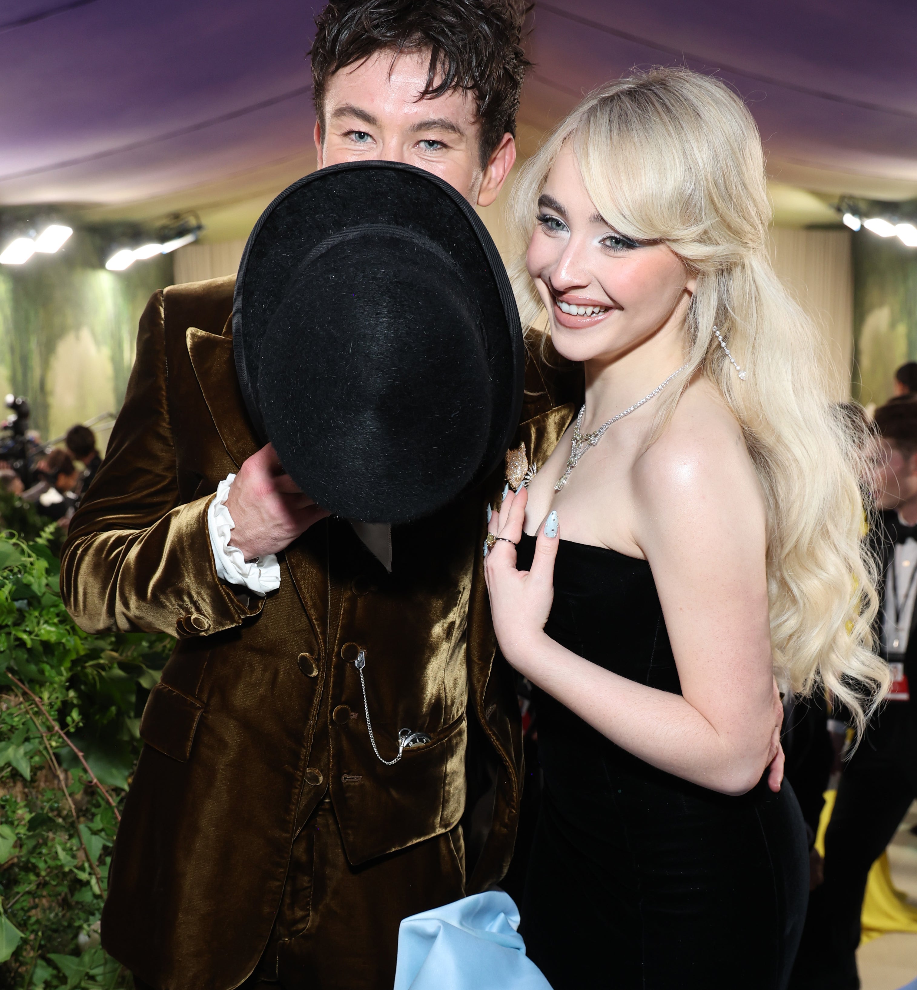 Barry Keoghan holding a hat partly covering his face, and Sabrina smiling, both dressed in elegant attire at a formal event