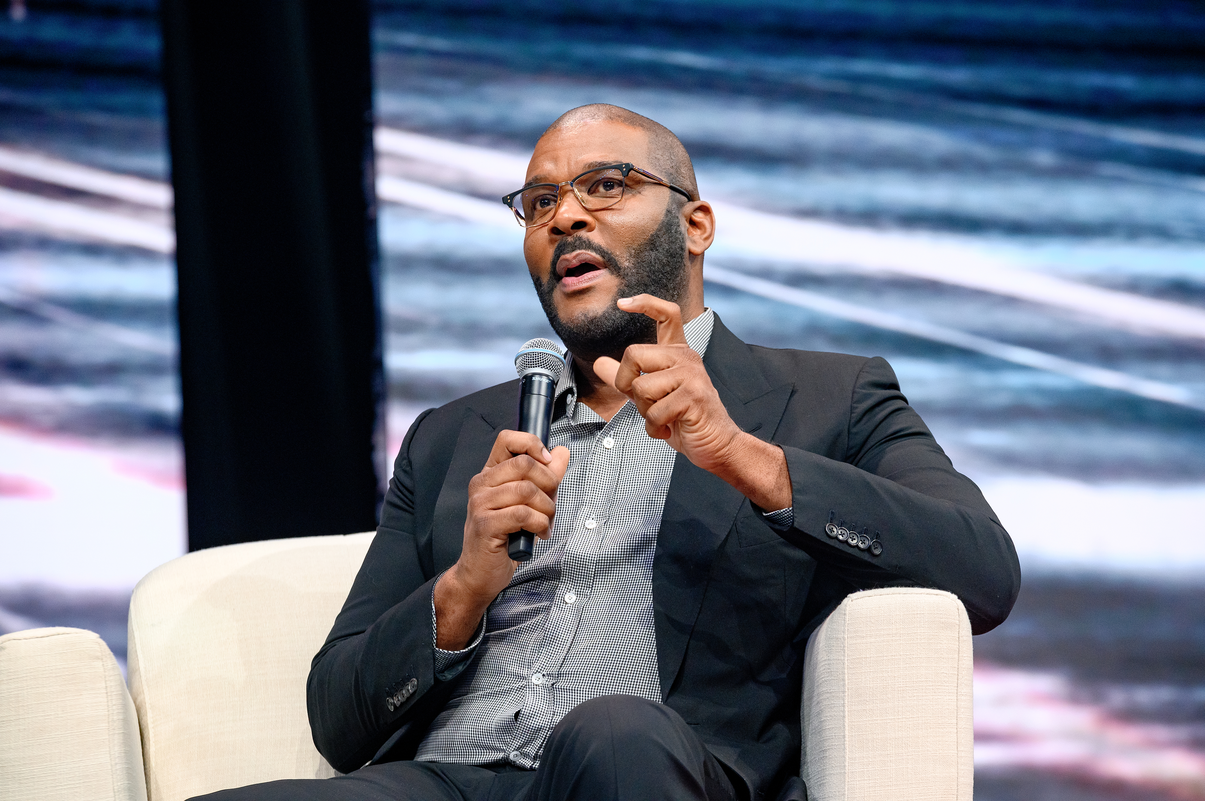 Tyler Perry sits on a stage holding a microphone, gesturing with his hand while speaking