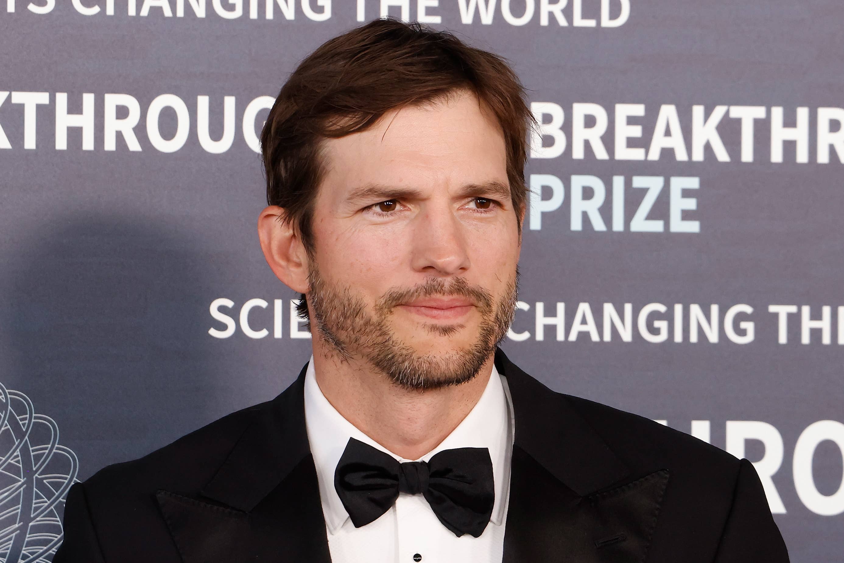 Ashton Kutcher in a tuxedo at the Breakthrough Prize event. Background text reads, "Changing the world," "Breakthrough Prize," and "Science changing the world."