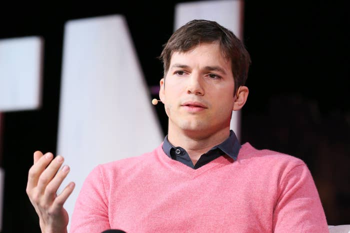 Ashton Kutcher speaking onstage, wearing a sweater over a collared shirt, with a small microphone attached to his face