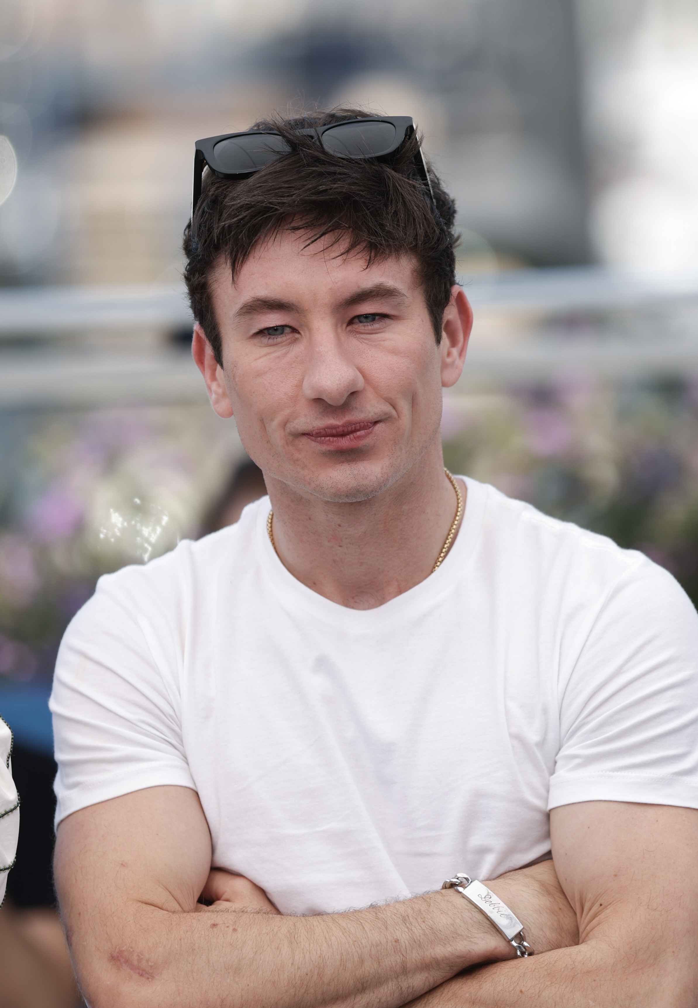 Barry Keoghan, with sunglasses resting on his head, smiles slightly while crossing his arms and wearing a casual white t-shirt