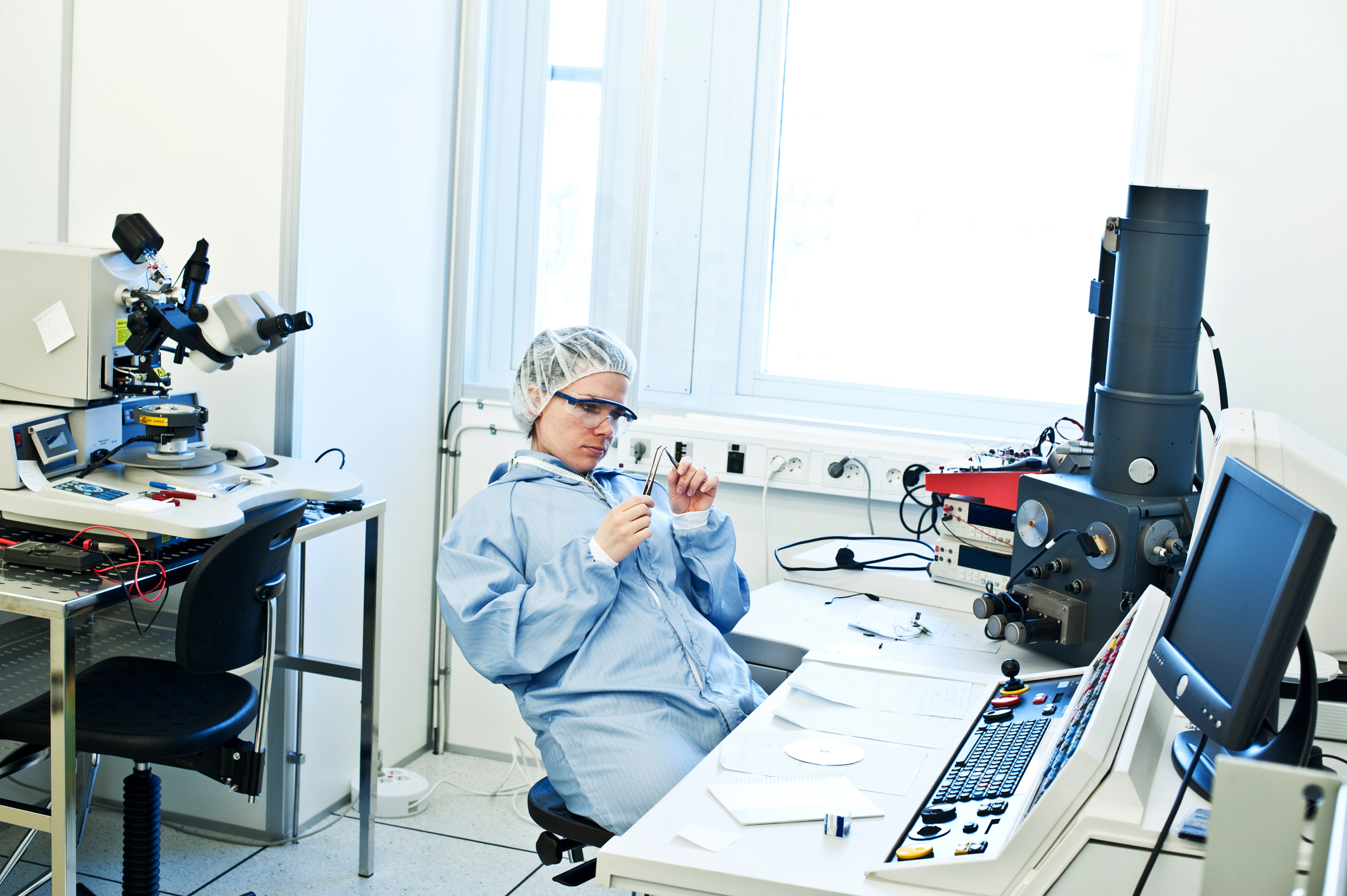 A person in protective lab clothing and goggles sits at a desk in a laboratory, holding lab tools, with various scientific equipment around them