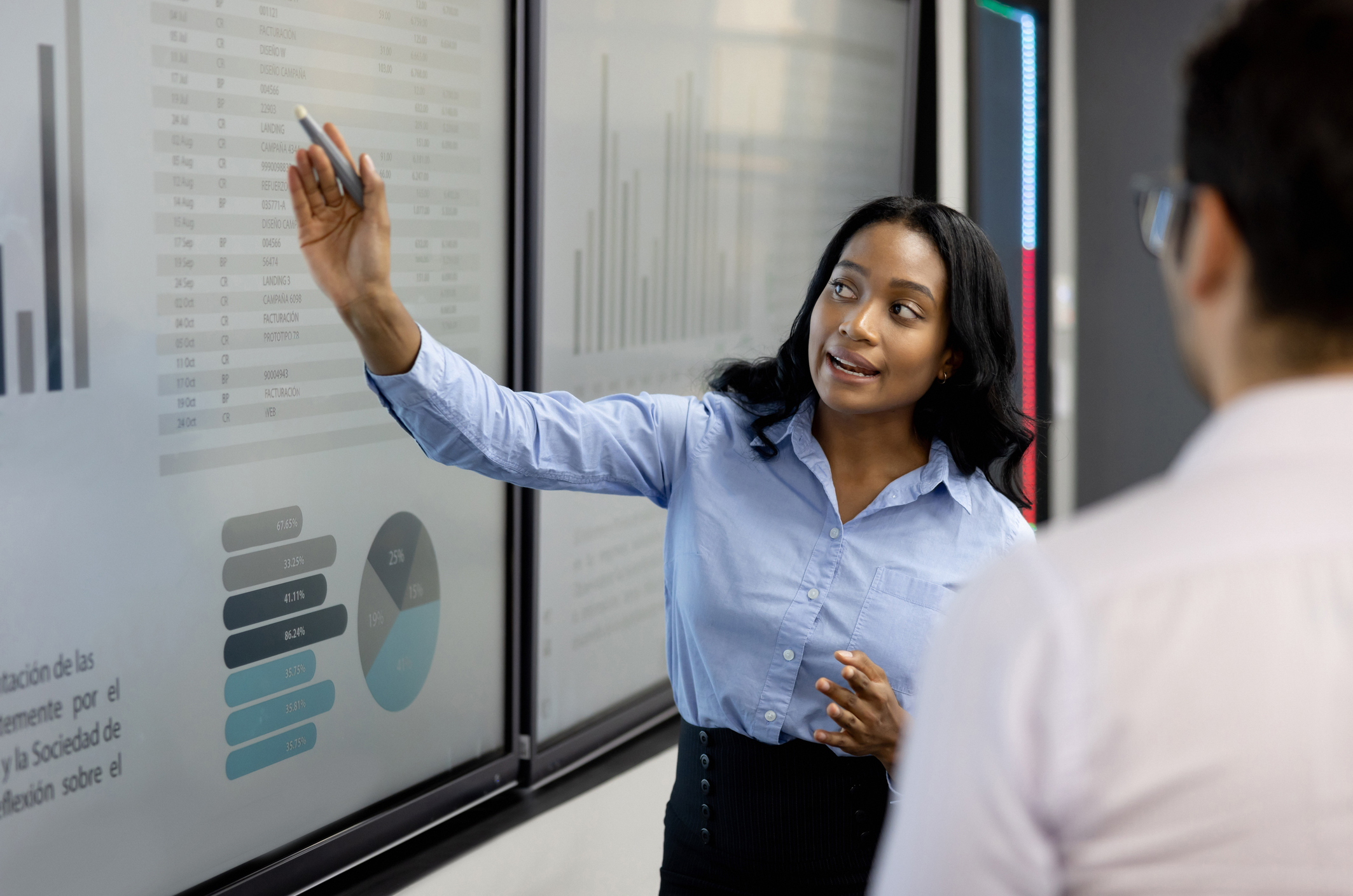 A woman in a business setting points at data charts on a screen, explaining to a colleague. She wears a light blouse and dark pants