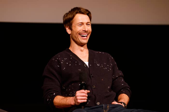 A man sits on stage, smiling and holding a microphone, wearing a casual black cardigan and jeans. The background is dark. The man's identity is unknown