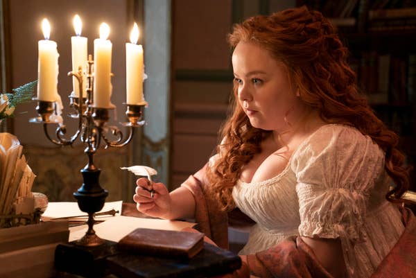 Nicola Coughlan is seated at a desk holding a quill pen next to a candle holder with lit candles in a scene from Bridgerton