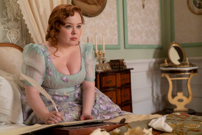 Nicola Coughlan, dressed in a period costume, sits at a table writing with a quill in a scene from Bridgerton