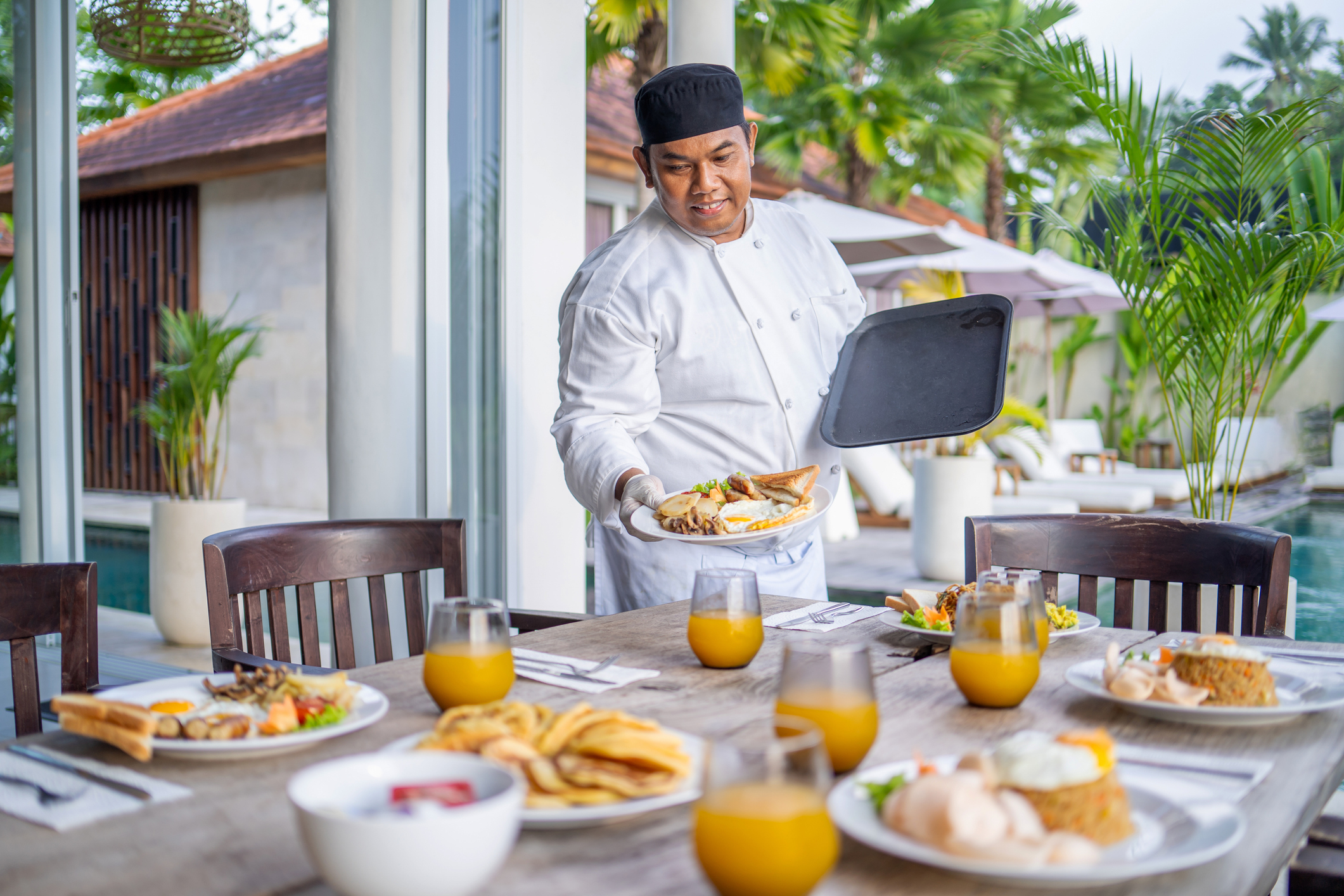A chef in a professional uniform serves food on an outdoor dining table near a pool. Several plates of breakfast items and glasses of juice are on the table