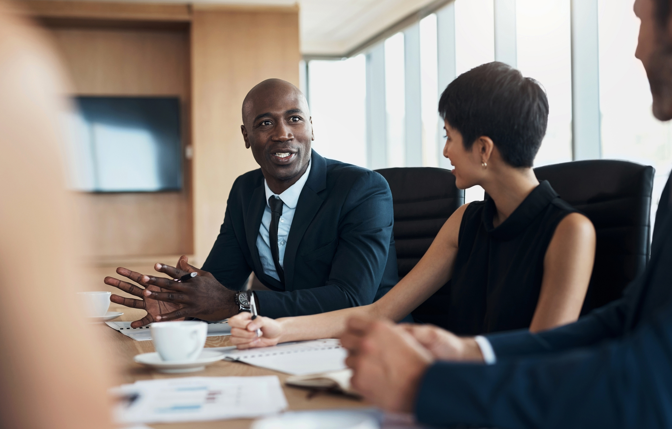 A group of business professionals in a meeting room, seated at a table with documents and coffee cups, engaged in a discussion. Unknown individuals