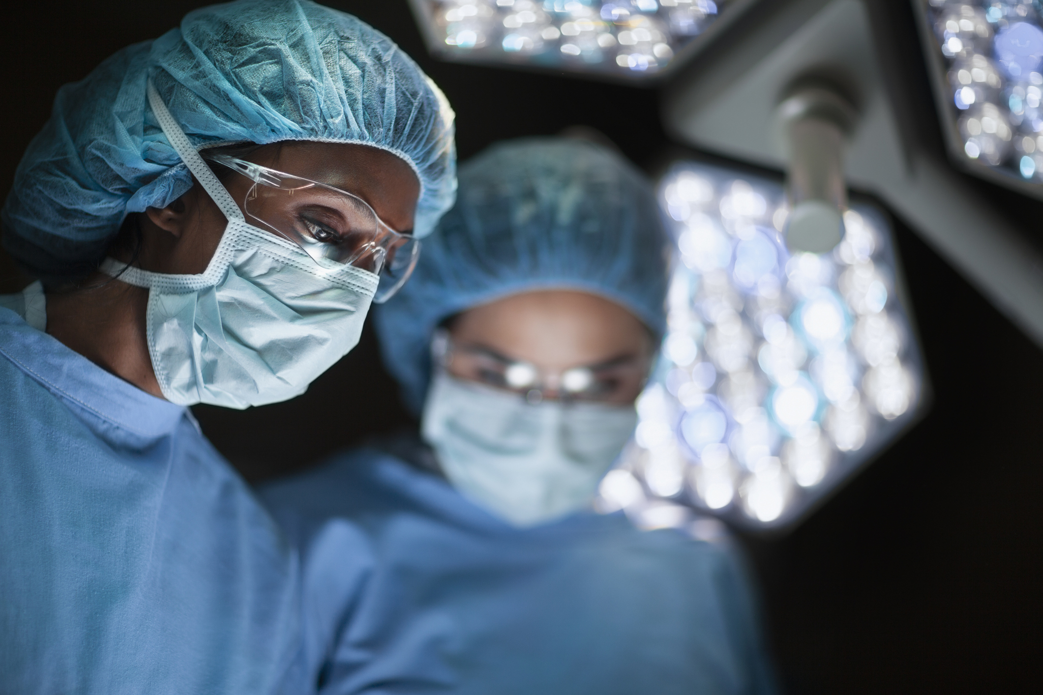 Two surgeons in surgical attire, including masks, goggles, and hairnets, focus intently on a patient under bright operating lights