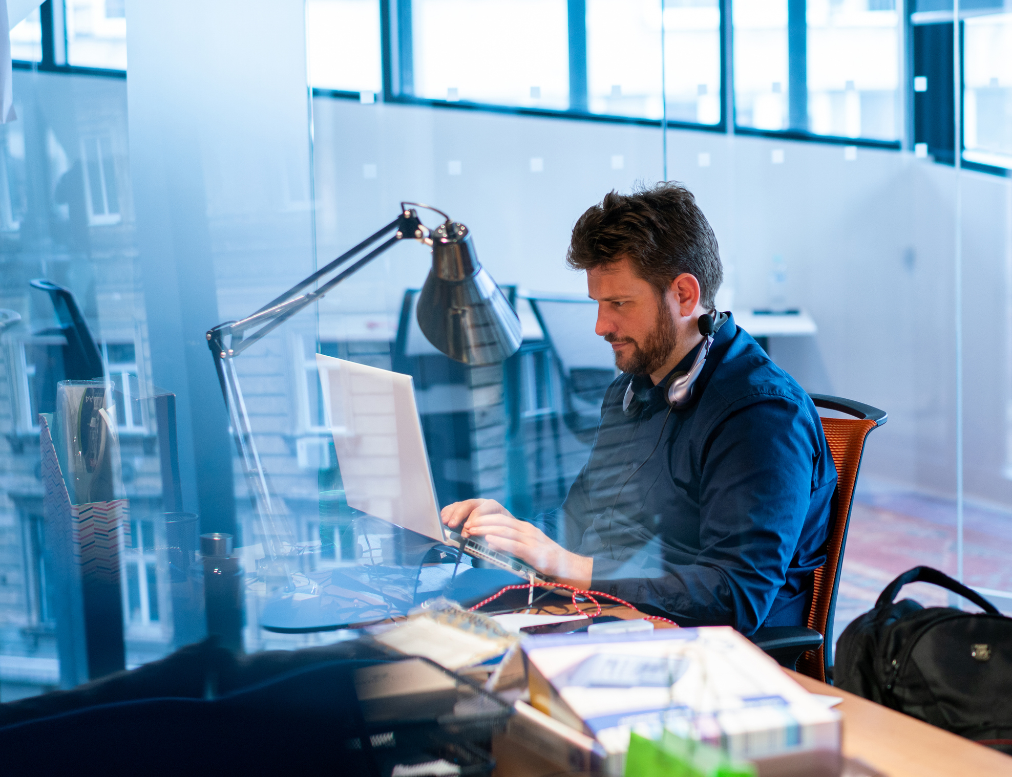 A man is seated at a desk in a modern office, focused on working on his laptop. He has headphones around his neck and is surrounded by work materials