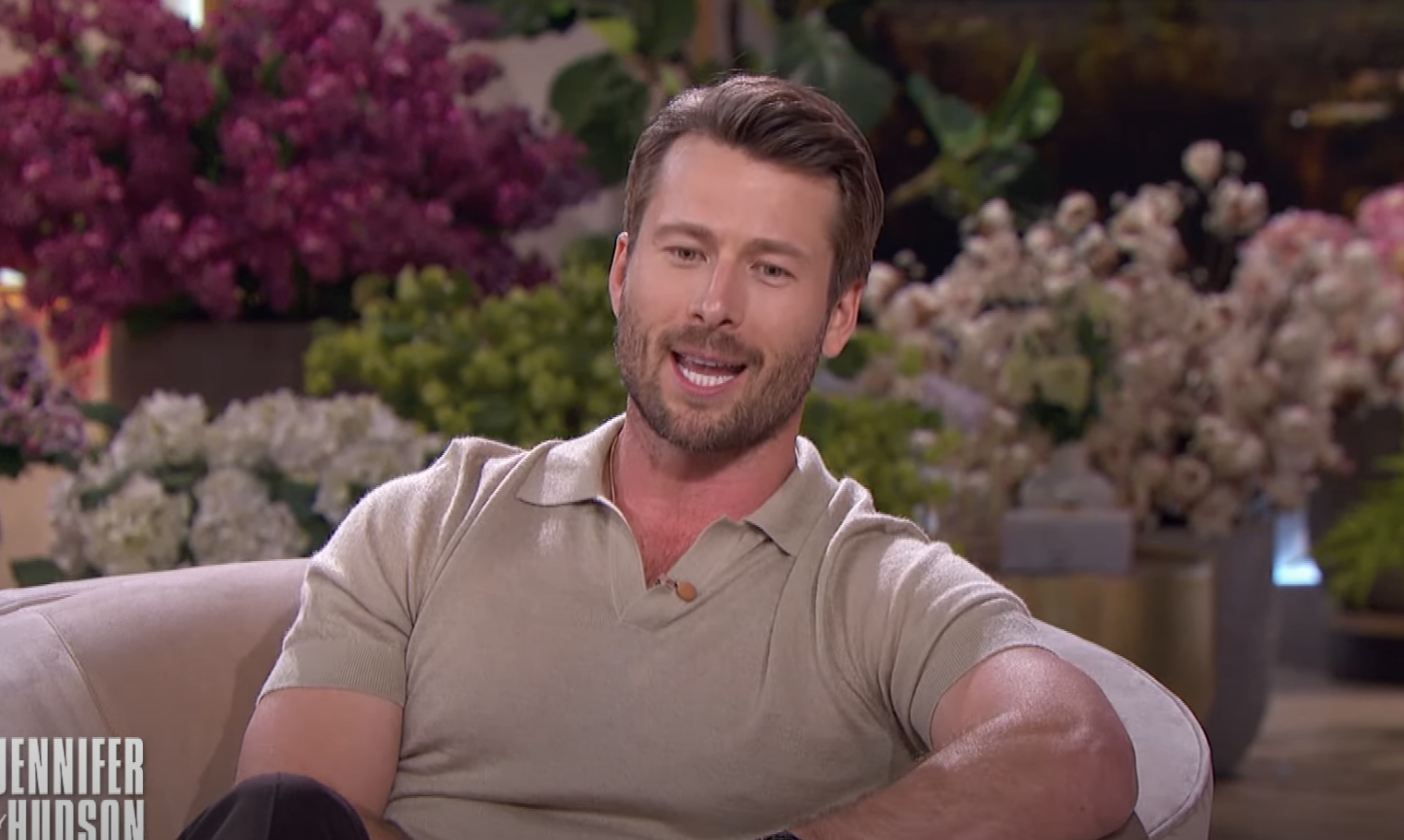 Glen Powell speaks during an interview on The Jennifer Hudson Show, seated in a relaxed setting with floral arrangements in the background