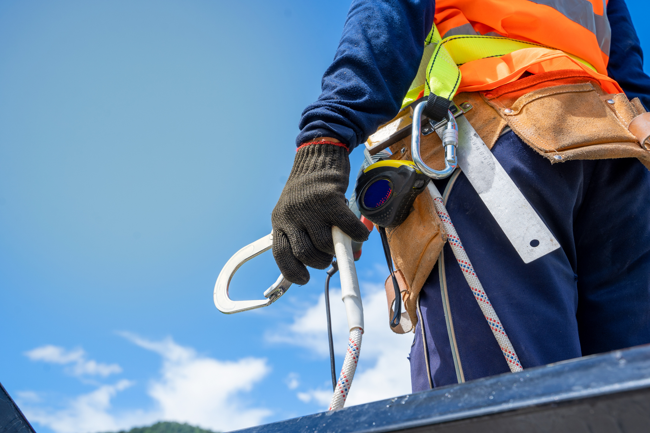 A construction worker in safety gear, holding a harness and rope, against a blue sky background