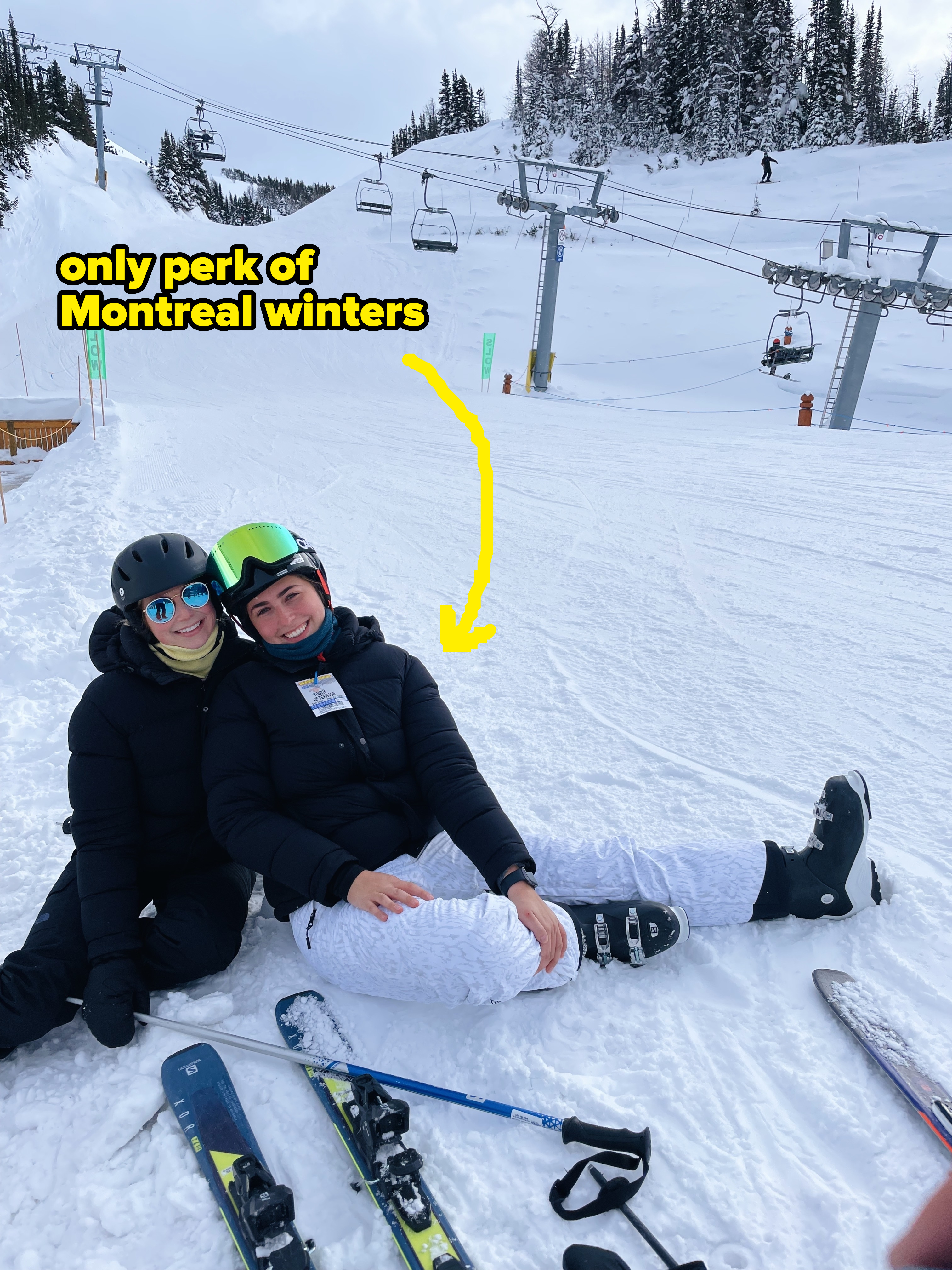 Two people sitting in the snow wearing ski gear, with ski poles and skis on the ground, in a ski resort with chairlifts and snow-covered trees in the background