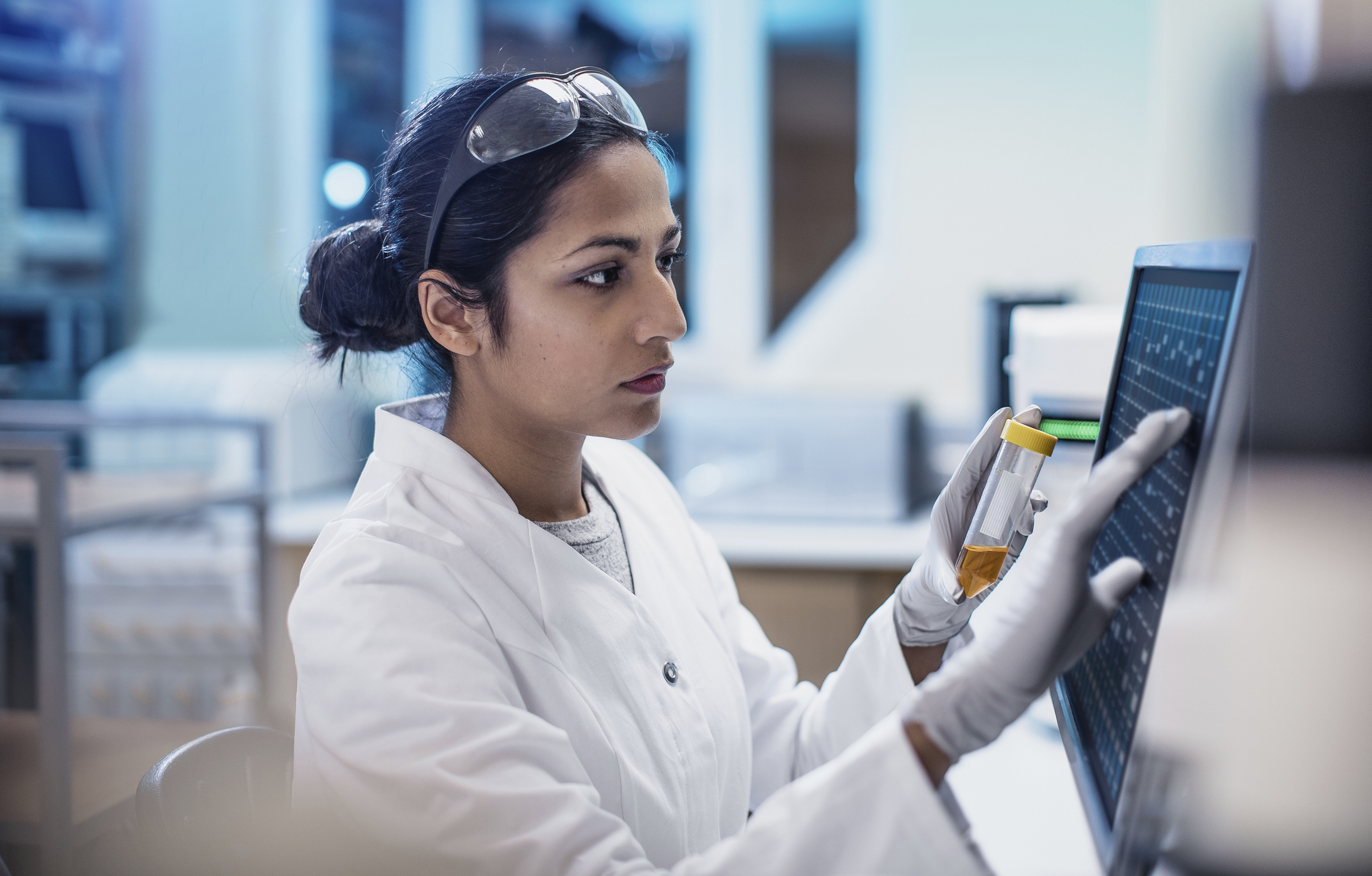 A scientist in a lab coat and gloves works on a computer while holding a test tube