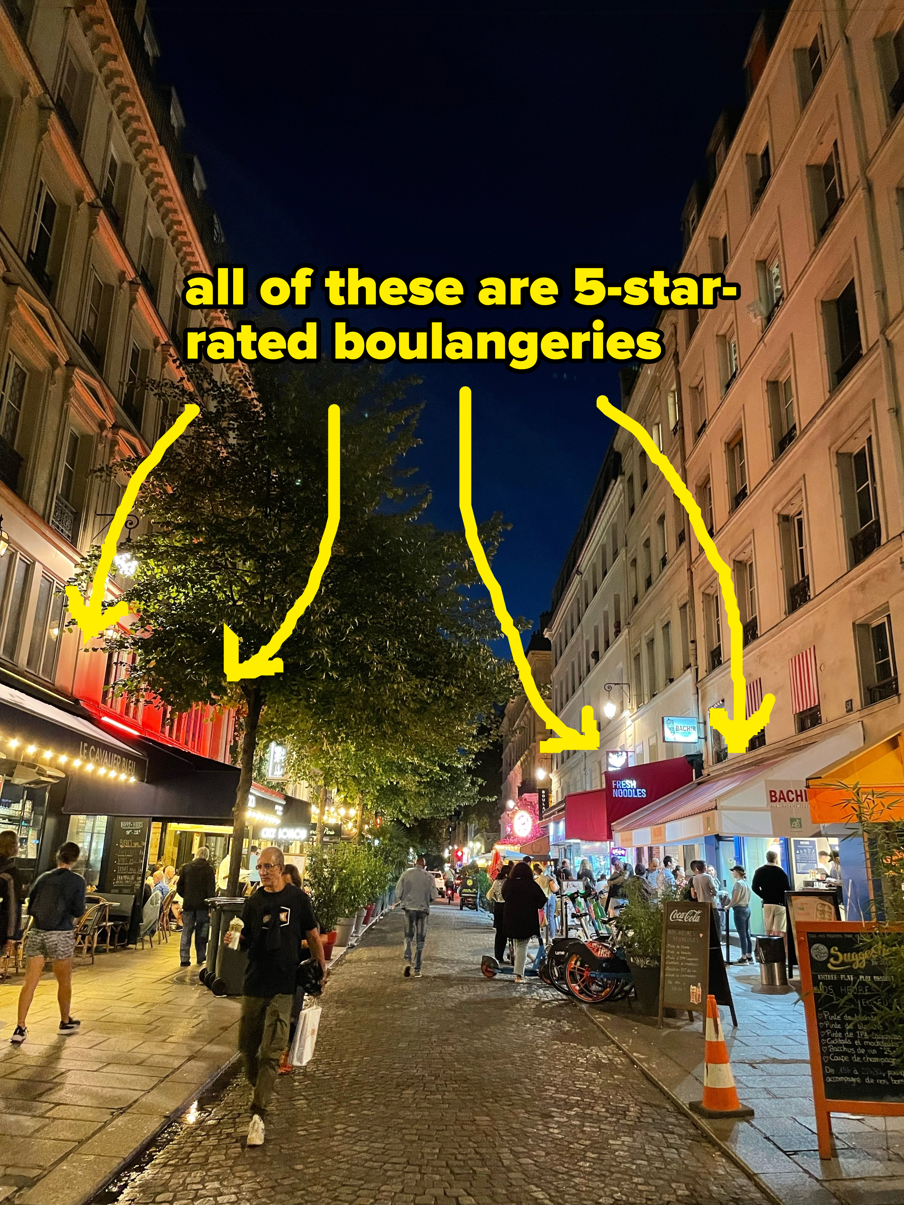 People stroll on a European street lined with cafes and buildings at night. Some are dining outside, while others walk or ride bicycles down the cobblestone path