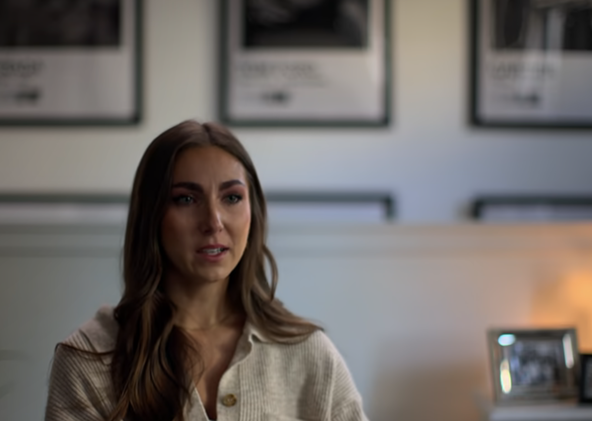 A woman with long hair sits indoors, speaking. Netflix trailer screengrab with black-and-white images on the wall behind her