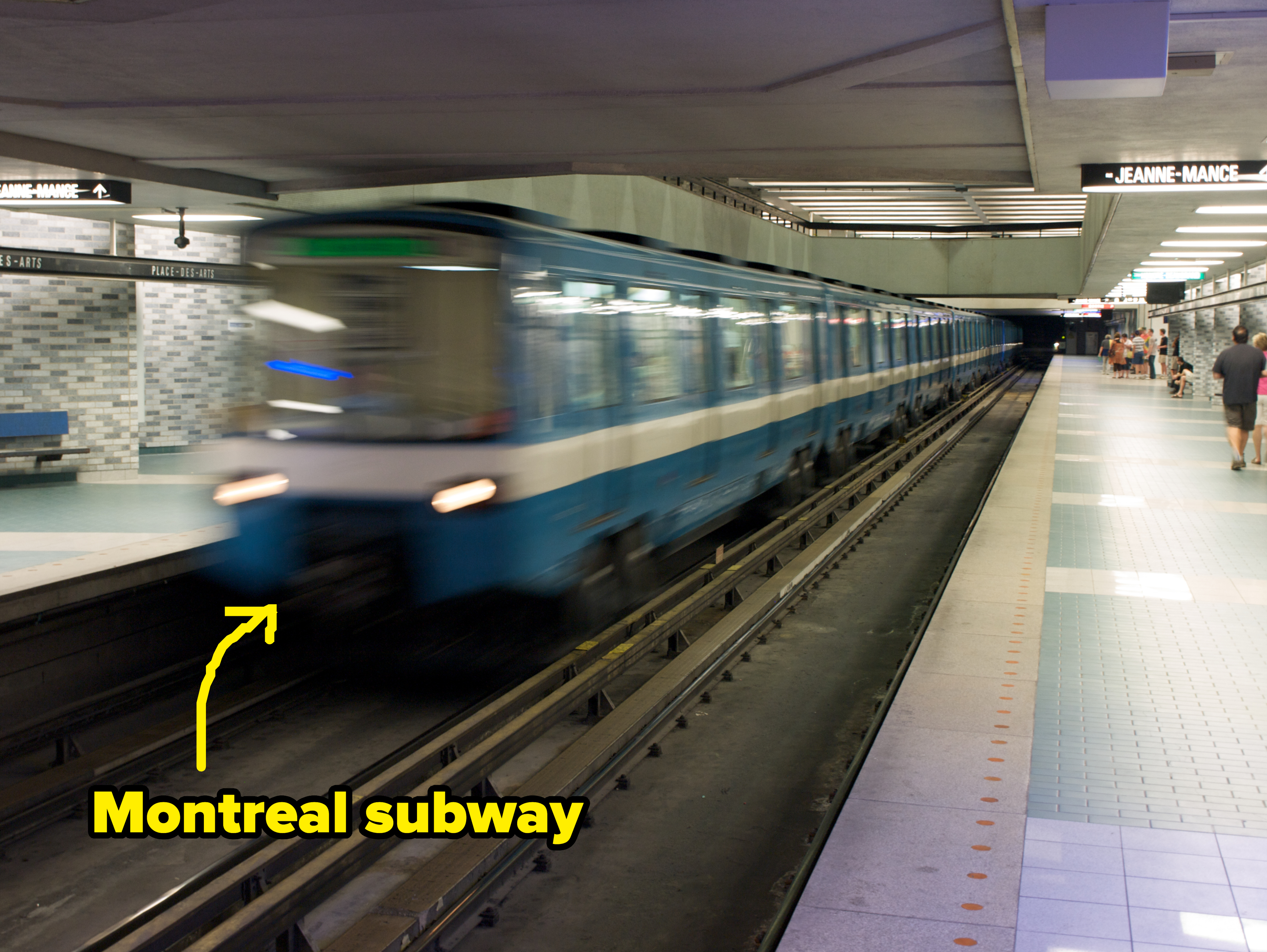 A subway train speeds through an underground station with visible signage. One person is walking on the platform