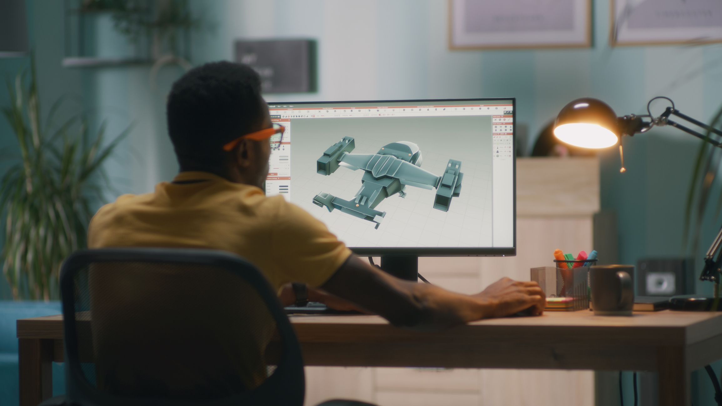 A person wearing glasses sits at a desk, using a computer to design a 3D model of a futuristic vehicle. The workspace features a plant and a lamp