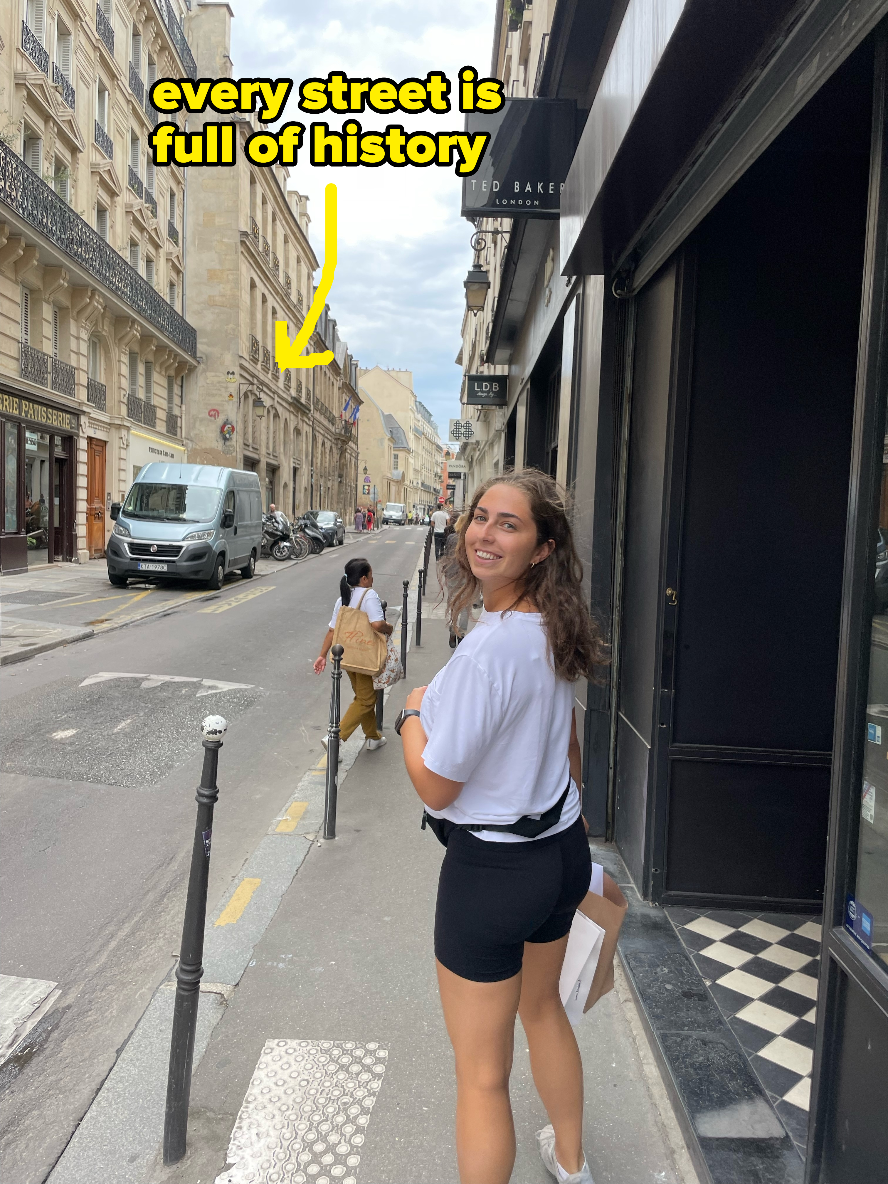 Woman in casual attire, holding a paper, smiles while walking on a European street lined with buildings and parked cars