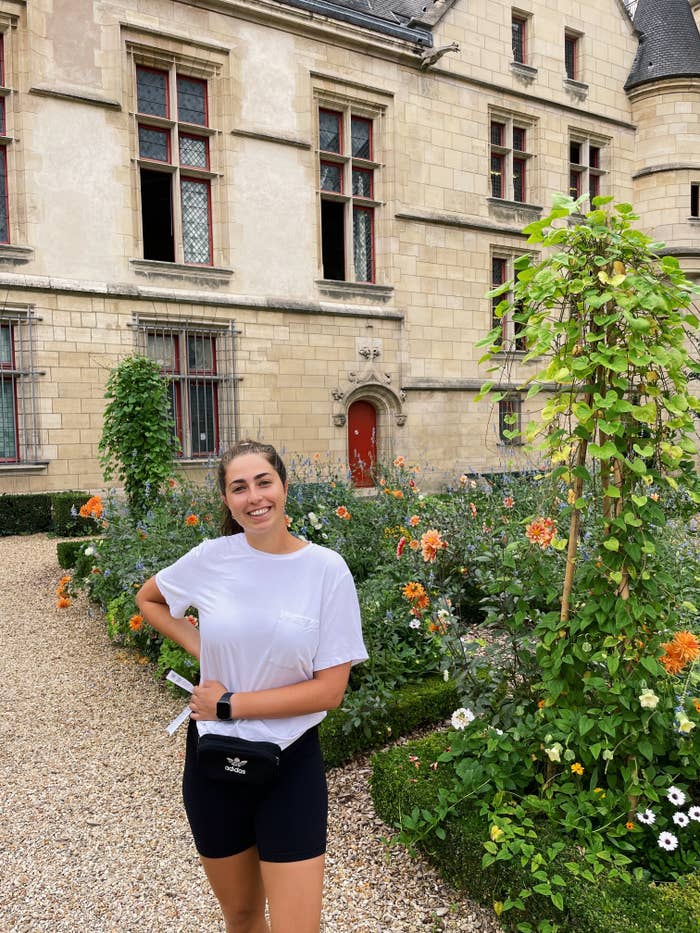 Person standing in front of a historic building with flowers in the garden, wearing a casual white t-shirt and black shorts. No known celebrities